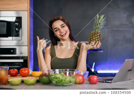Healthy food at home. Happy woman is showing pineapple in modern kitchen. Vegan and vegeterian lifestile. Healthy food at home. Happy woman is showing pineapple in modern kitchen. Vegan and vegeterian lifestile. 108354500