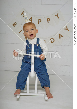 First birthday baby boy party. Happy beautiful baby of European appearance with white hair and blue eyes on white background. First birthday baby boy party. Happy beautiful baby of European appearance with white hair and blue eyes on white background. 108354509
