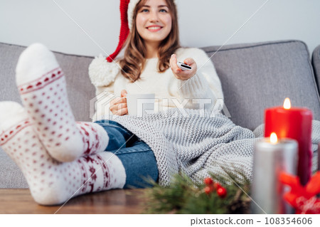 Hand holding remote controller with blurred smiling woman in santa hat, warm socks with cup of hot drink sitting on comfortable couch watching movie, TV with christmas decoration mood. Cozy winter Hand holding remote controller with blurred smiling woman in santa hat, warm socks with cup of hot drink sitting on comfortable couch watching movie, TV with christmas decoration mood. Cozy winter 108354606