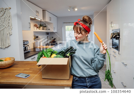 Smiling woman eating fresh carrot from her healthy food delivery box. Box with vegetables and phone with Veganuary app on the kitchen table. Start of a healthy life concept. Online home food delivery. 108354640