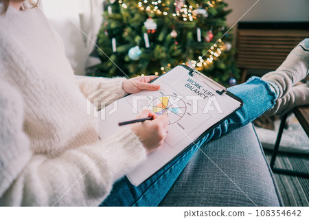 Woman drawing work-life balance wheel sitting on the sofa at home. Self-reflection and life planning for next new year. Personal Year review. Coaching tools. Finding Balance in Life. Selective focus. 108354642