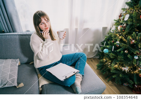 Young Woman with cup is hand writing New year's goals on note paper in new year day. New year resolutions list. Goals, resolutions, plans, action, checklist concept. New Year template, copy space Young Woman with cup is hand writing New year's goals on note paper in new year day. New year resolutions list. Goals, resolutions, plans, action, checklist concept. New Year template, copy space 108354648