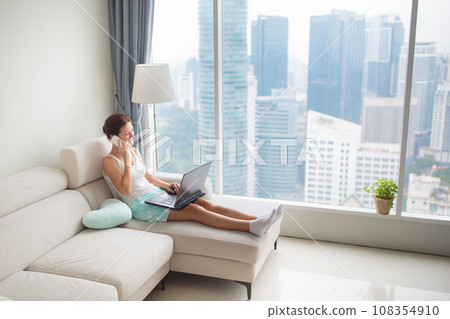 Woman working at laptop at big window with city view Woman working at laptop at big window with city view 108354910