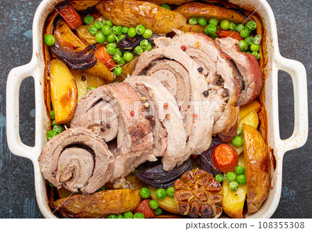 Rolled sliced pork roasted in white casserole dish with potatoes, vegetables and herbs on blue dark concrete rustic background top view. Baked pork roll with vegetables for dinner Rolled sliced pork roasted in white casserole dish with potatoes, vegetables and herbs on blue dark concrete rustic background top view. Baked pork roll with vegetables for dinner 108355308