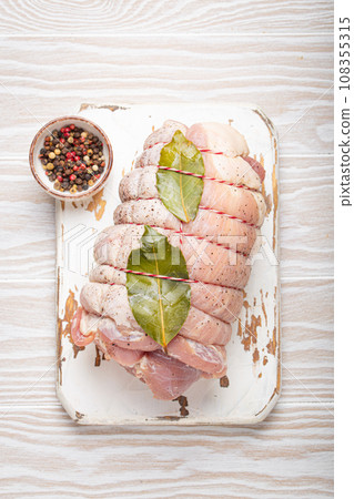 Raw wrapped rolled sliced pork with bay leaf and seasonings on white cutting board on rustic white wooden background top view. Pork roll ready to be prepared 108355315
