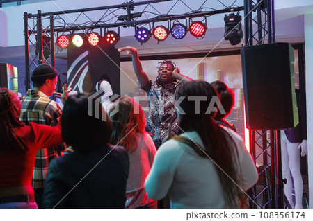 People clubbing and dancing at electronic music concert in nightclub. Crowd partying on dancefloor while african american dj performing electronic music on stage with lights in club 108356174