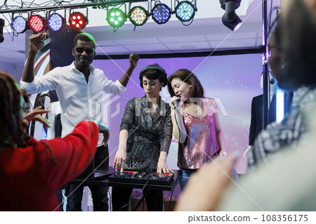 Electronic music band performing on stage with spotlights at concert in nightclub. Dj and singer musicians playing while crowd cheering on dancefloor and partying in club Electronic music band performing on stage with spotlights at concert in nightclub. Dj and singer musicians playing while crowd cheering on dancefloor and partying in club 108356175