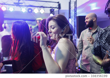 Young woman partying and dancing in crowded nightclub while attending social gathering event. Clubber girl moving to live music rhythm on dancefloor surrounded with people Young woman partying and dancing in crowded nightclub while attending social gathering event. Clubber girl moving to live music rhythm on dancefloor surrounded with people 108356199