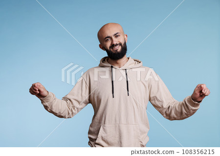 Smiling arab man pretending to hold banner in two hands and looking at camera with cheerful facial expression. Young adult joyful person advertising product studio portrait Smiling arab man pretending to hold banner in two hands and looking at camera with cheerful facial expression. Young adult joyful person advertising product studio portrait 108356215