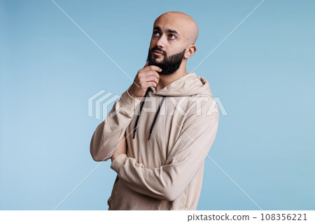 Young arab man touching chin thoughtfully and looking away with doubt facial expression. Person wearing casual clothes while thinking and solving problem with pensive emotions 108356221