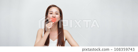 Woman in love, holding a little paper heart over eye, standing over white background, panorama Woman in love, holding a little paper heart over eye, standing over white background, panorama 108359743