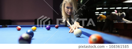 Focused woman hits white ball leaning above blue pool table Focused woman hits white ball leaning above blue pool table 108361585