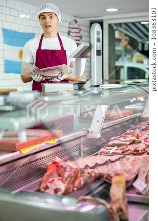 Positive young salesman demonstrating piece of meat in butcher shop 108363101