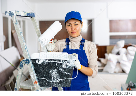 Woman painter in overalls with paint roller in building under construction Woman painter in overalls with paint roller in building under construction 108363372