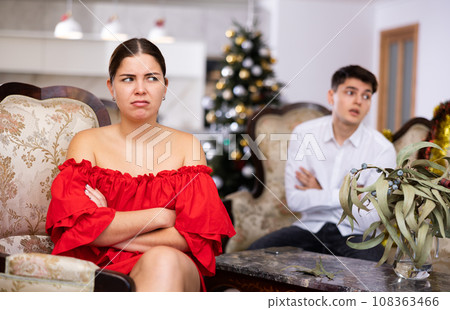 Displeased young European man and woman in red dress yelling at each other on sofa during preparations for New Year in living room Displeased young European man and woman in red dress yelling at each other on sofa during preparations for New Year in living room 108363466