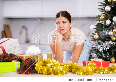 Disappointed young European woman sad about for New year preparations next to the Christmas tree and wrapped gifts in living room 108363558