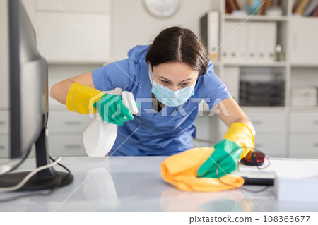 Woman in protective mask wipes dust from table in office 108363677