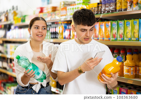Young man and woman searching on internet contents of soft drinks together in supermarket Young man and woman searching on internet contents of soft drinks together in supermarket 108363680