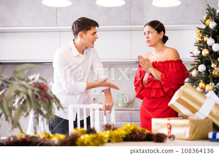 Upset young couple arguing among themselves before X-mas Eve near Christmas tree in kitchen at home Upset young couple arguing among themselves before X-mas Eve near Christmas tree in kitchen at home 108364136