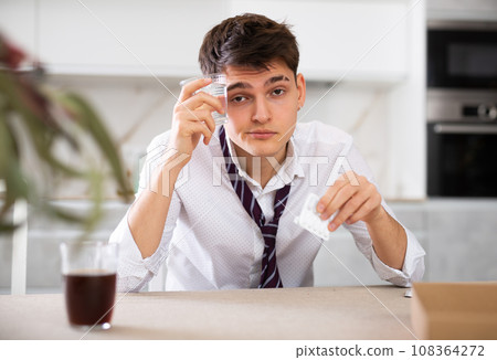 Sick young man with hangover drinks pills while sitting at table 108364272