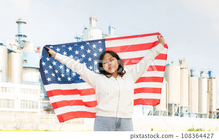 Happy asian girl with flag of usa standing in front of industrial scenery Happy asian girl with flag of usa standing in front of industrial scenery 108364427