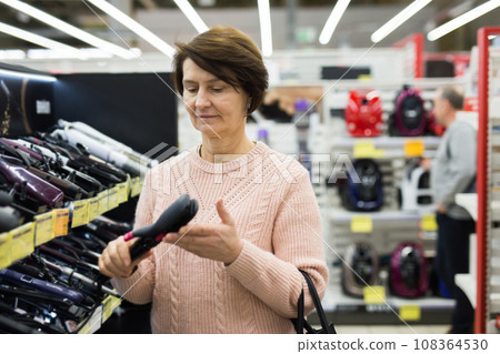 Middle aged woman choosing hair iron in appliance shop Middle aged woman choosing hair iron in appliance shop 108364530