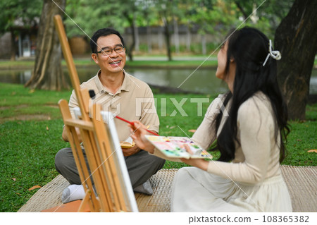 Happy 50s man and young adult daughter painting picture on the green grass at park 108365382