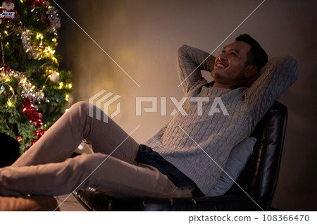 Happy Asian man in a cosy sweater is relaxing on a chair in a cosy living room on a Christmas night. 108366470