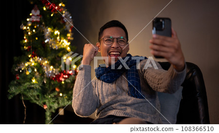 A cheerful Asian man is watching a sports match on his phone and enjoying his Christmas night alone A cheerful Asian man is watching a sports match on his phone and enjoying his Christmas night alone 108366510