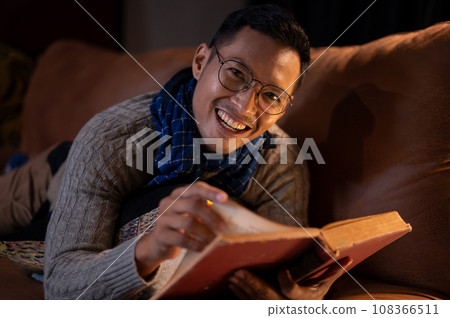 A happy Asian man in cosy clothes is reading a book while lying on sofa in his living room at night. A happy Asian man in cosy clothes is reading a book while lying on sofa in his living room at night. 108366511