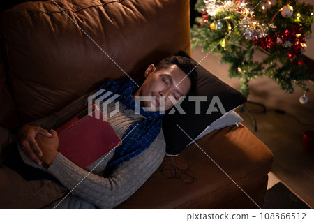 A tired Asian man falls asleep while reading a book, sleeping on a sofa in the living room. A tired Asian man falls asleep while reading a book, sleeping on a sofa in the living room. 108366512