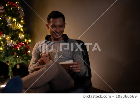 A handsome and happy Asian man is reading a book while sipping coffee on a Christmas night. A handsome and happy Asian man is reading a book while sipping coffee on a Christmas night. 108366569