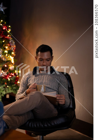 A focused Asian man is reading a book while sipping coffee on a chair near by a Christmas tree. A focused Asian man is reading a book while sipping coffee on a chair near by a Christmas tree. 108366570