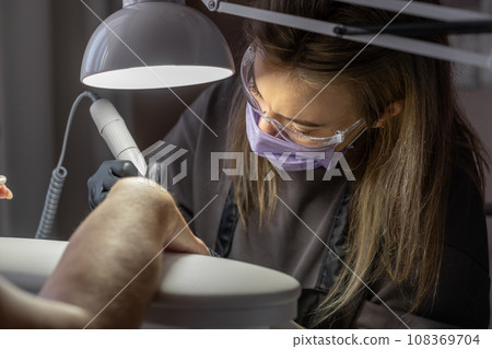 Manicurist at work. Professional manicurist removes old worn nail design from nails of client using modern electric drill. Preparing fingernails for making fresh manicure and applying new nail design 108369704