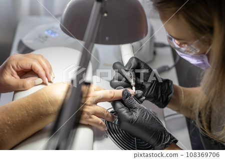 Manicurist at work. Professional manicurist removes old worn nail design from nails of client using modern electric drill. Preparing fingernails for making fresh manicure and applying new nail design 108369706