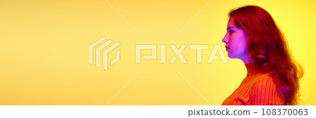 Side view portrait of young redhaired girl looking straight against yellow studio background in neon light 108370063