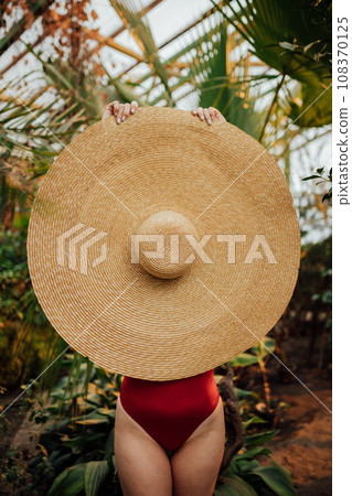 Woman in red swimsuit hides his face behind hat standing among tropical plants in greenhouse Woman in red swimsuit hides his face behind hat standing among tropical plants in greenhouse 108370125