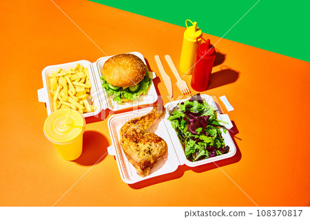 Opened paper box with salad with baked chicken, hamburger with fried potato, sauces and drinks over orange-green background. Concept of fast food, delivery. Opened paper box with salad with baked chicken, hamburger with fried potato, sauces and drinks over orange-green background. Concept of fast food, delivery. 108370817