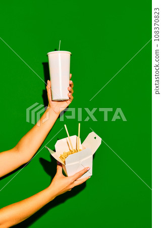 Take it away. Close up image of hands holds one opened paper box with noodles and refreshing drink against green background. Concept of fast food, delivery. Take it away. Close up image of hands holds one opened paper box with noodles and refreshing drink against green background. Concept of fast food, delivery. 108370823