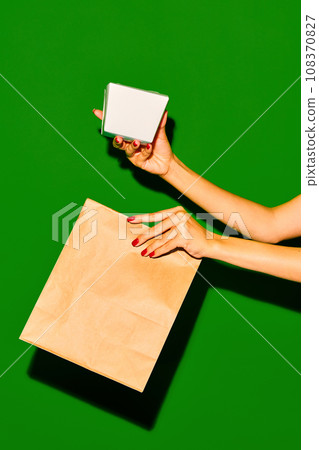 Take it away. Close up image of two female hands holding paper box and bag against vivid green background. Concept of fast food, delivery. 108370827