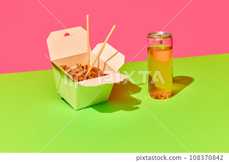 Opened WOK paper box of Asian noodles fast-food with bottle of refreshing drink against green-pink background. Concept of fast food, delivery. Opened WOK paper box of Asian noodles fast-food with bottle of refreshing drink against green-pink background. Concept of fast food, delivery. 108370842