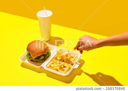 Unpacking tasting fast-food. Unrecognizable woman eat hamburger with soda and fried potato in paper boxes against vivid yellow background. Concept of delivery. Unpacking tasting fast-food. Unrecognizable woman eat hamburger with soda and fried potato in paper boxes against vivid yellow background. Concept of delivery. 108370848