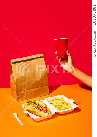 Tasty hotdog with sausage served mustard and ketchup with green salad with refreshing drinks and fried potato against vivid red background. Concept of fast food, delivery. 108370861