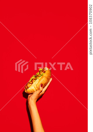 Hand holding hot tasty American hotdog with beef sausage, sauces with green salad against vivid red background. Concept of fast food, delivery. Hand holding hot tasty American hotdog with beef sausage, sauces with green salad against vivid red background. Concept of fast food, delivery. 108370902