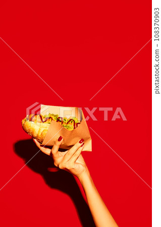 Two hands holding hot tasty traditional American hotdog with beef sausage, sauces with green salad against vivid red background. Concept of fast food, delivery. Two hands holding hot tasty traditional American hotdog with beef sausage, sauces with green salad against vivid red background. Concept of fast food, delivery. 108370903