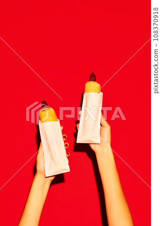 Two hands holding hot delicious traditional American hotdog with beef sausage and sauces against vivid red background. Concept of fast food, delivery. Two hands holding hot delicious traditional American hotdog with beef sausage and sauces against vivid red background. Concept of fast food, delivery. 108370918