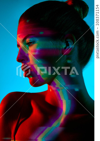 Portrait of beautiful young woman against blue background with multicolored neon reflection on face Portrait of beautiful young woman against blue background with multicolored neon reflection on face 108371154