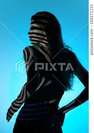 Back view of elegant young woman in jeans, looking away, posing against blue background in neon light. Stripes of shadow Back view of elegant young woman in jeans, looking away, posing against blue background in neon light. Stripes of shadow 108371155
