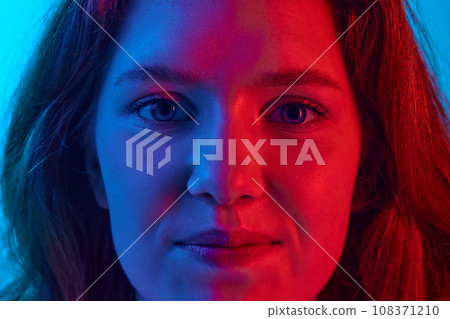 Close-up of young girl looking at camera with little smile against blue studio background in neon light 108371210