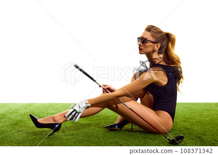 Attractive, beautiful, elegant young woman with slim tanned body, in bodysuit and heels posing on grass with golf club isolated over white background 108371342